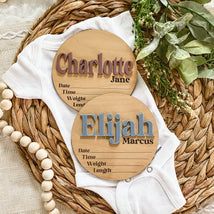Personalized wooden baby name signs for Charlotte Jane and Elijah Marcus on a woven mat with baby onesie.