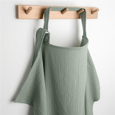 Muslin nursing cover in fern green hanging on a wooden hook, featuring adjustable straps for breastfeeding privacy.