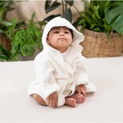 Toddler in a white hooded bathrobe sitting on a light surface, with green plants in the background.