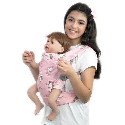 A girl wearing a pink toy carrier featuring unicorns, holding a doll that resembles an infant.