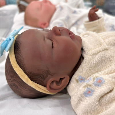 Reborn baby doll with African American features, wearing a yellow headband and cream-colored outfit with floral design.