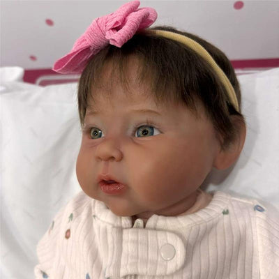 Reborn baby doll with dark brown hair, a pink bow, and wearing a white outfit with flower patterns.