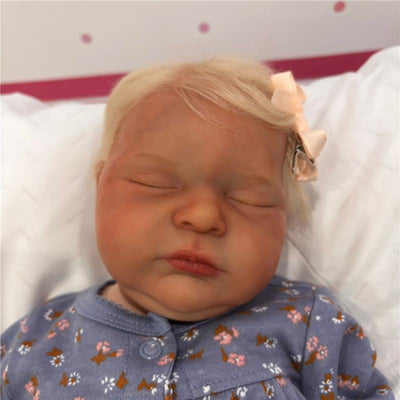 Reborn baby doll with blonde hair and a pink bow, wearing a floral-patterned blue outfit, eyes closed.