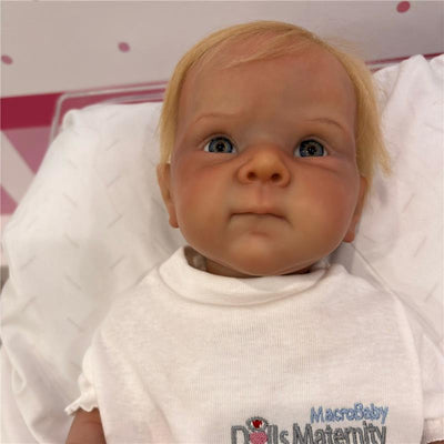 Reborn baby doll named Bettie, featuring white vinyl, rooted blonde hair, and blue eyes, wearing a white shirt.