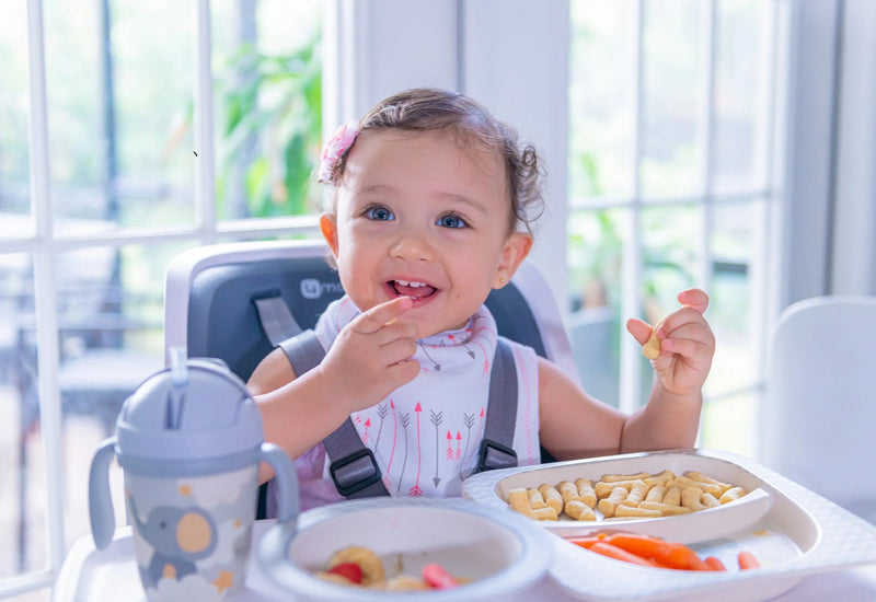 Happy baby eating snacks in high chair with sippy cup and divided plate, perfect for baby mealtime essentials