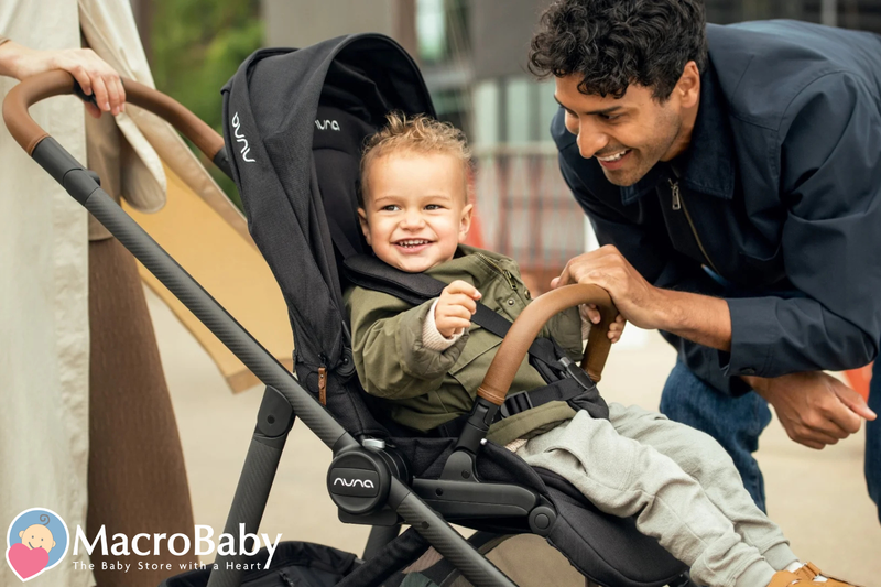Happy baby in black Nuna stroller with dad smiling nearby, outdoor family moment