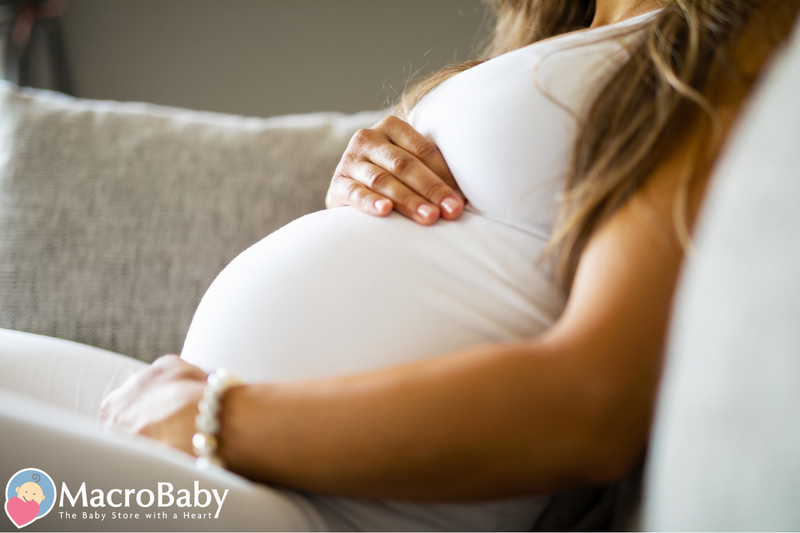 Pregnant woman in white dress gently holding her belly, sitting on a couch
