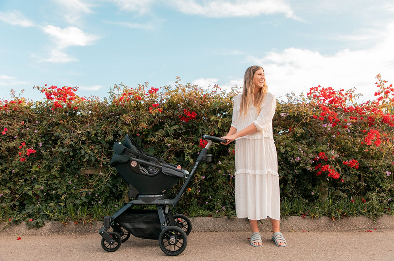 Mom pushing baby stroller outdoors near red flowering hedge, enjoying sunny day with infant car seat attached