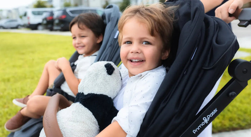 Happy toddler smiling in black double stroller with panda plush, perfect for baby gear and outdoor adventures.