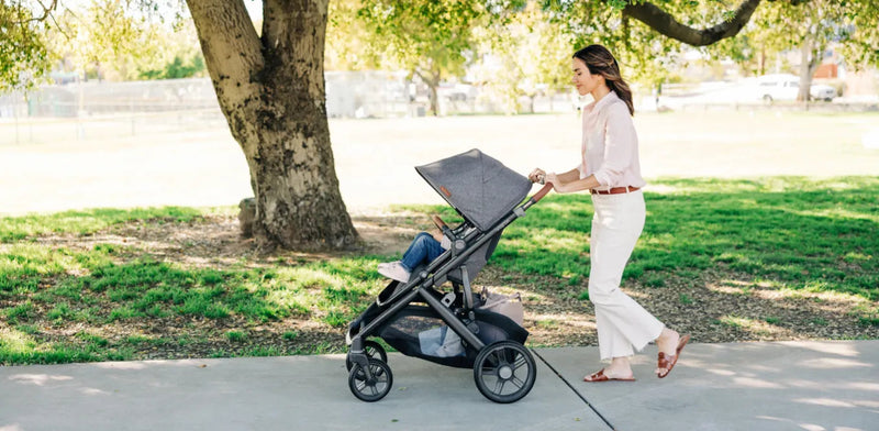 Woman pushing baby stroller in park with green grass and trees