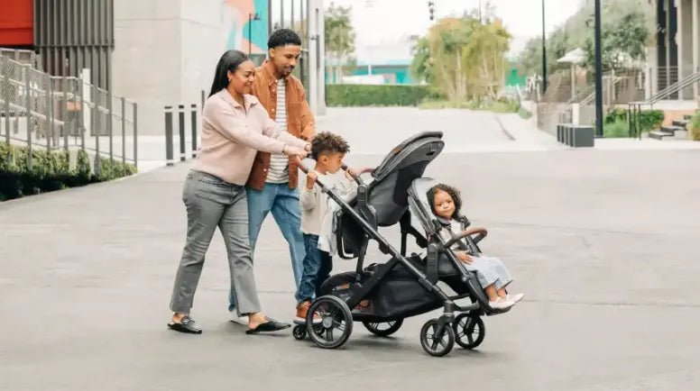Family walking outdoors with a double stroller, one child seated, one standing beside