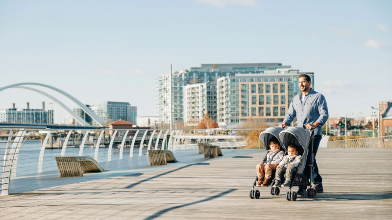 Dad pushing double stroller with two toddlers on waterfront boardwalk near city skyline
