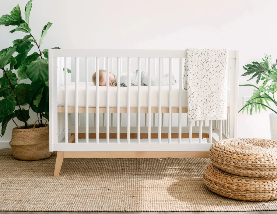 Baby sleeping in white modern crib with speckled blanket, natural rug, and potted plants in nursery
