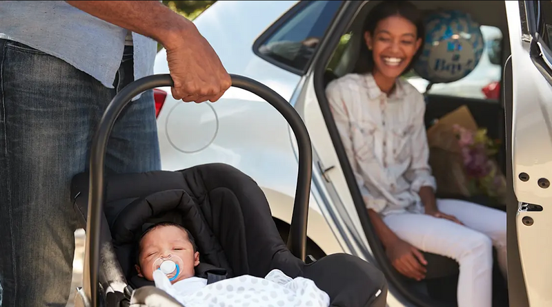 Newborn in infant car seat with pacifier, parent holding carrier near car, mom smiling inside