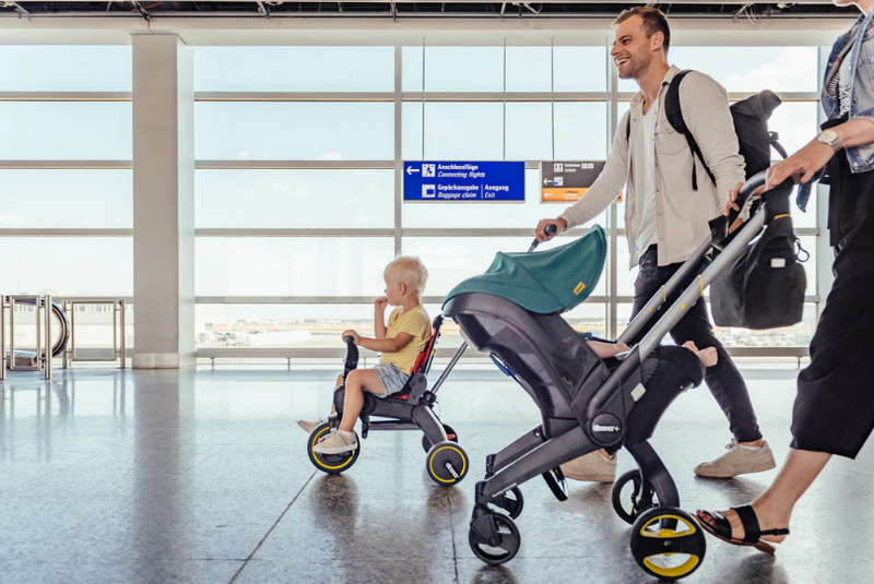 Dad pushing stroller with toddler riding tricycle at airport, ideal for travel with baby gear