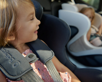 Toddler in black car seat with harness, smiling beside another child in light gray car seat