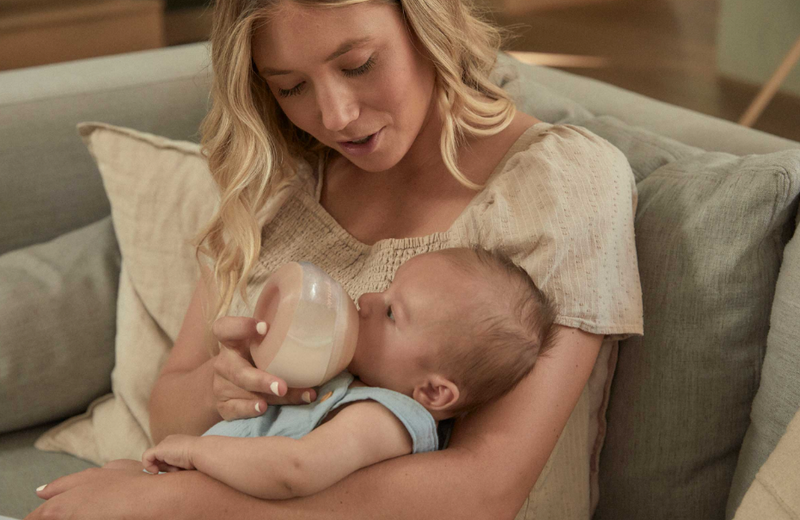 Mom feeding baby with bottle on cozy couch, baby essentials for newborn care