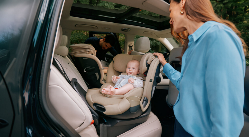 Baby in beige car seat inside SUV, mom adjusting harness, dad loading stroller in back