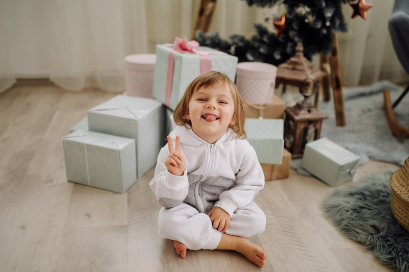 Happy toddler in white and gray onesie making peace sign surrounded by holiday gifts near Christmas tree