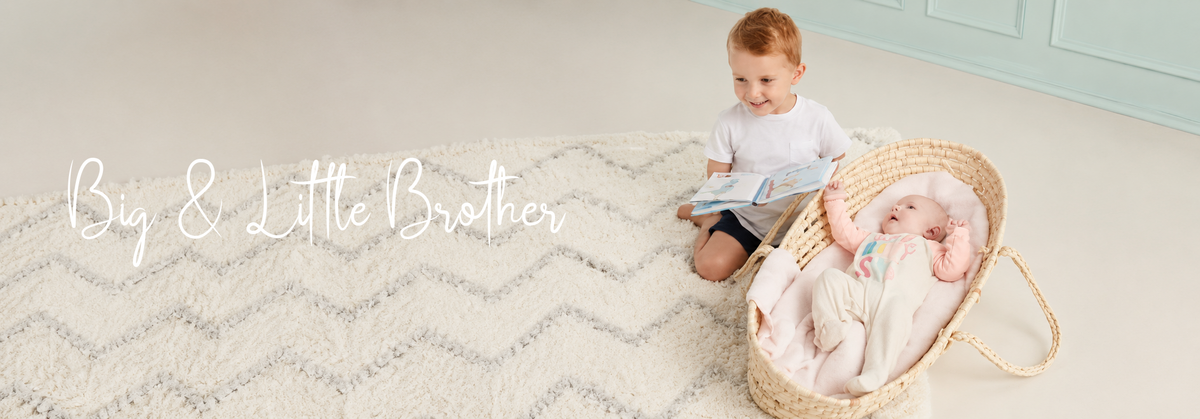 Big brother reading to baby in woven bassinet on chevron rug, cozy nursery scene