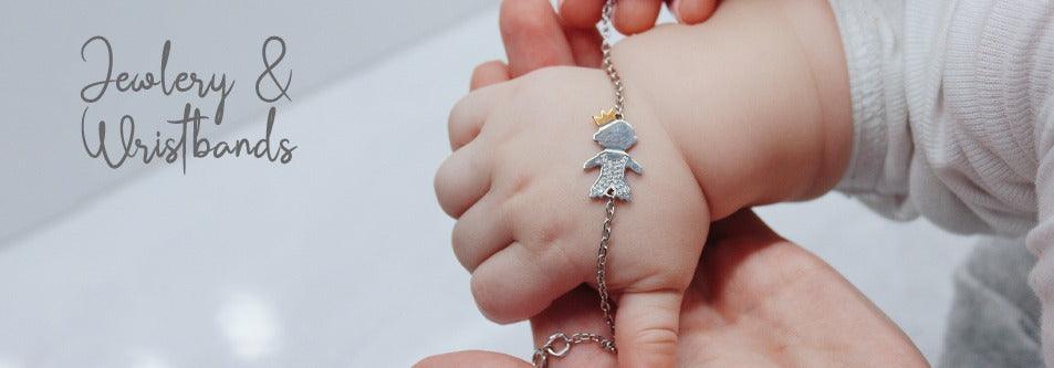 Baby wearing delicate silver crown charm bracelet, soft white outfit, gentle hand holding wrist