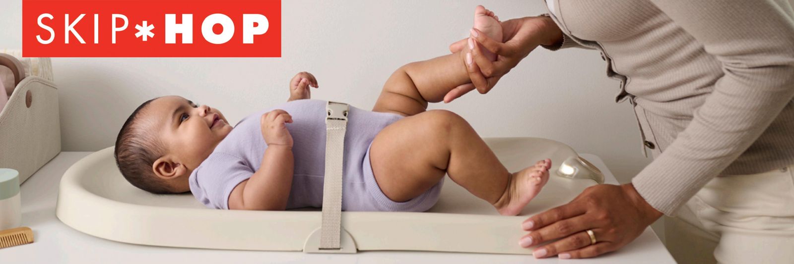 Baby on Skip Hop changing pad with safety strap, mom gently holding baby's legs during diaper change