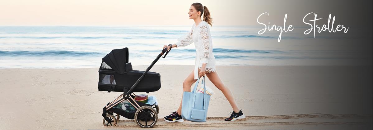 Woman pushing black single stroller on beach, carrying blue diaper bag, ocean backdrop