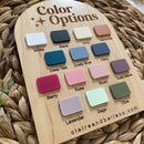 Color options swatch board with 14 pastel and neutral shades for baby products, displayed on woven mat