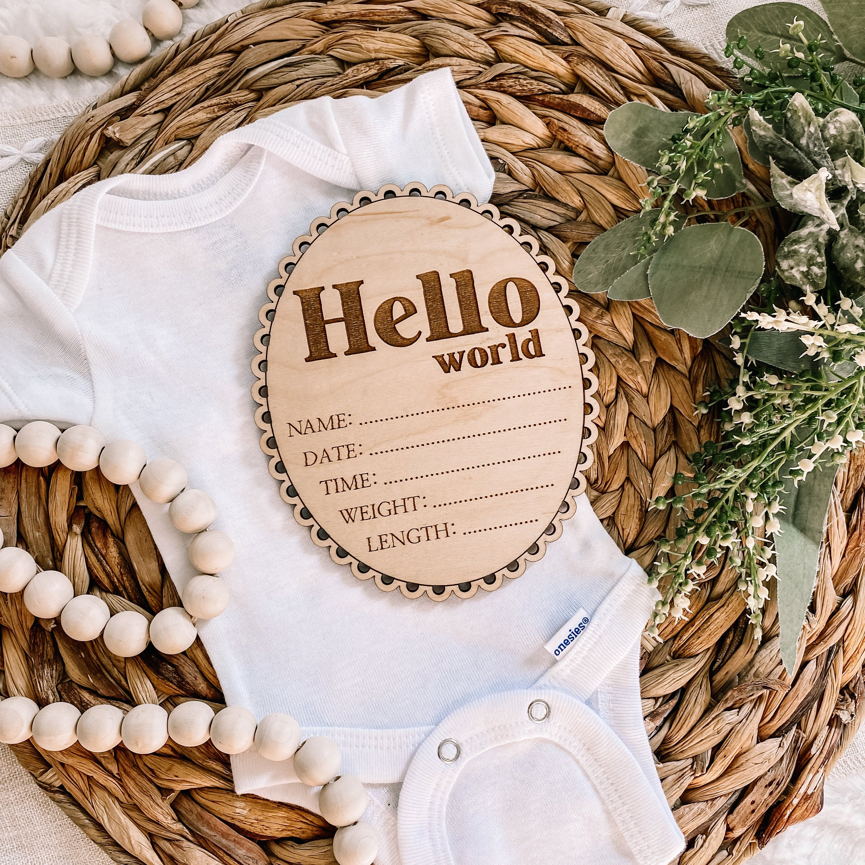 Hello World baby announcement sign with onesie, wooden beads, and greenery on woven mat
