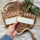 Hello My Name Is name tags with pink and purple lettering, held over woven mat and greenery