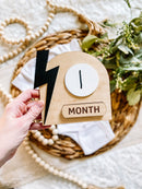 Wooden baby milestone sign with lightning bolt, marking first month, styled with baby clothes and greenery
