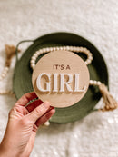 Wooden 'It's a Girl' sign for baby gender reveal, held over green basket with beads