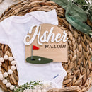 Personalized baby onesie with golf-themed name sign for Asher William, styled on woven mat