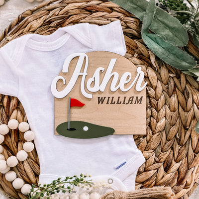 Personalized baby onesie with golf-themed name sign for Asher William, styled on woven mat