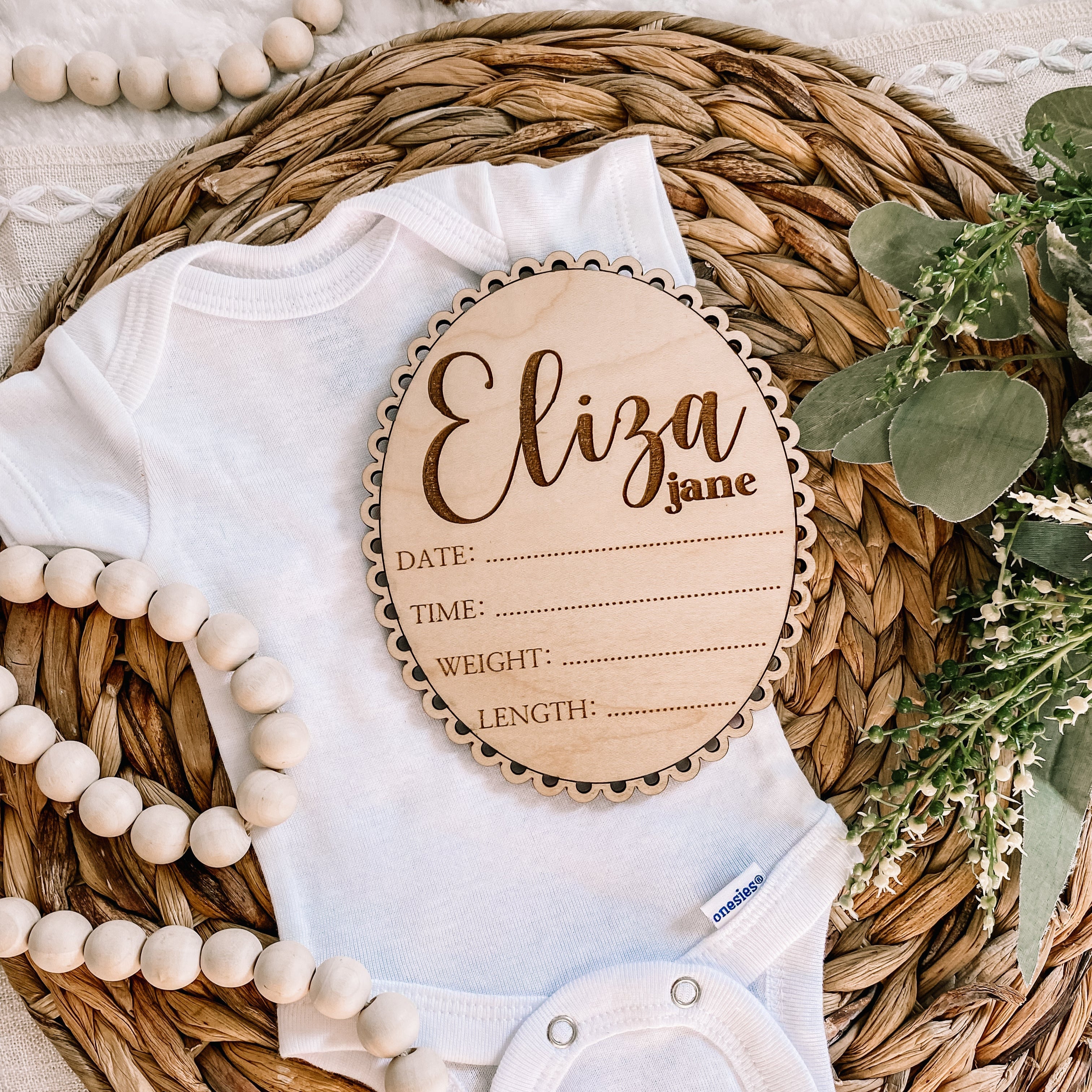 Personalized wooden baby milestone plaque with name Eliza Jane, placed on a white onesie with beads and greenery.