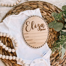 Personalized wooden baby milestone plaque with name Eliza Jane, placed on a white onesie with beads and greenery.