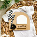 White baby onesie with checkered flag and 1-month milestone card on woven mat