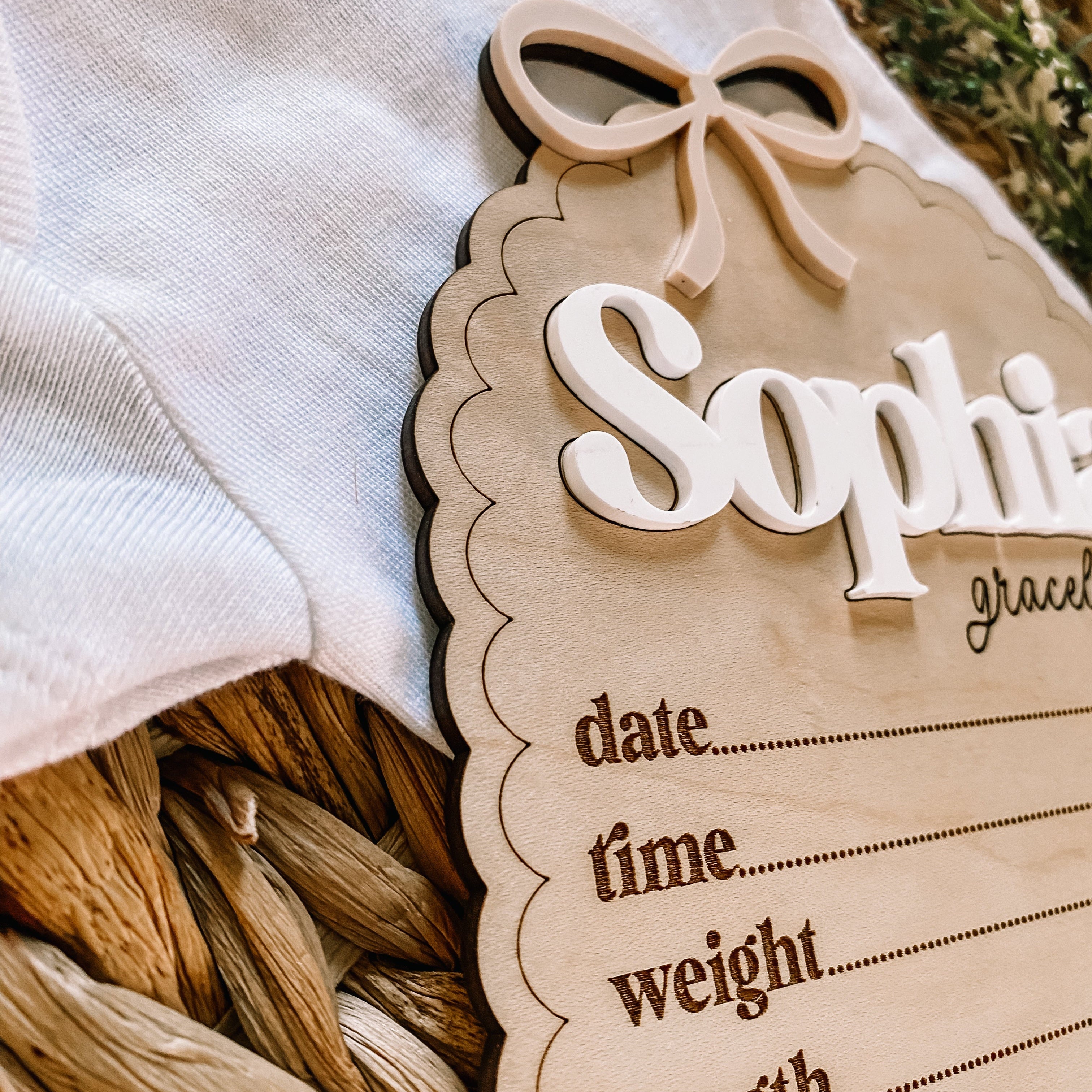 Personalized baby name sign with bow, nestled beside soft baby clothing in a wicker basket.