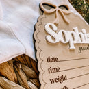 Personalized baby name sign with bow, nestled beside soft baby clothing in a wicker basket.