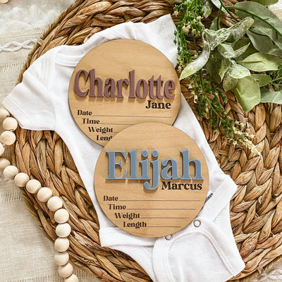Personalized wooden baby name signs for Charlotte Jane and Elijah Marcus on a woven mat with baby onesie.