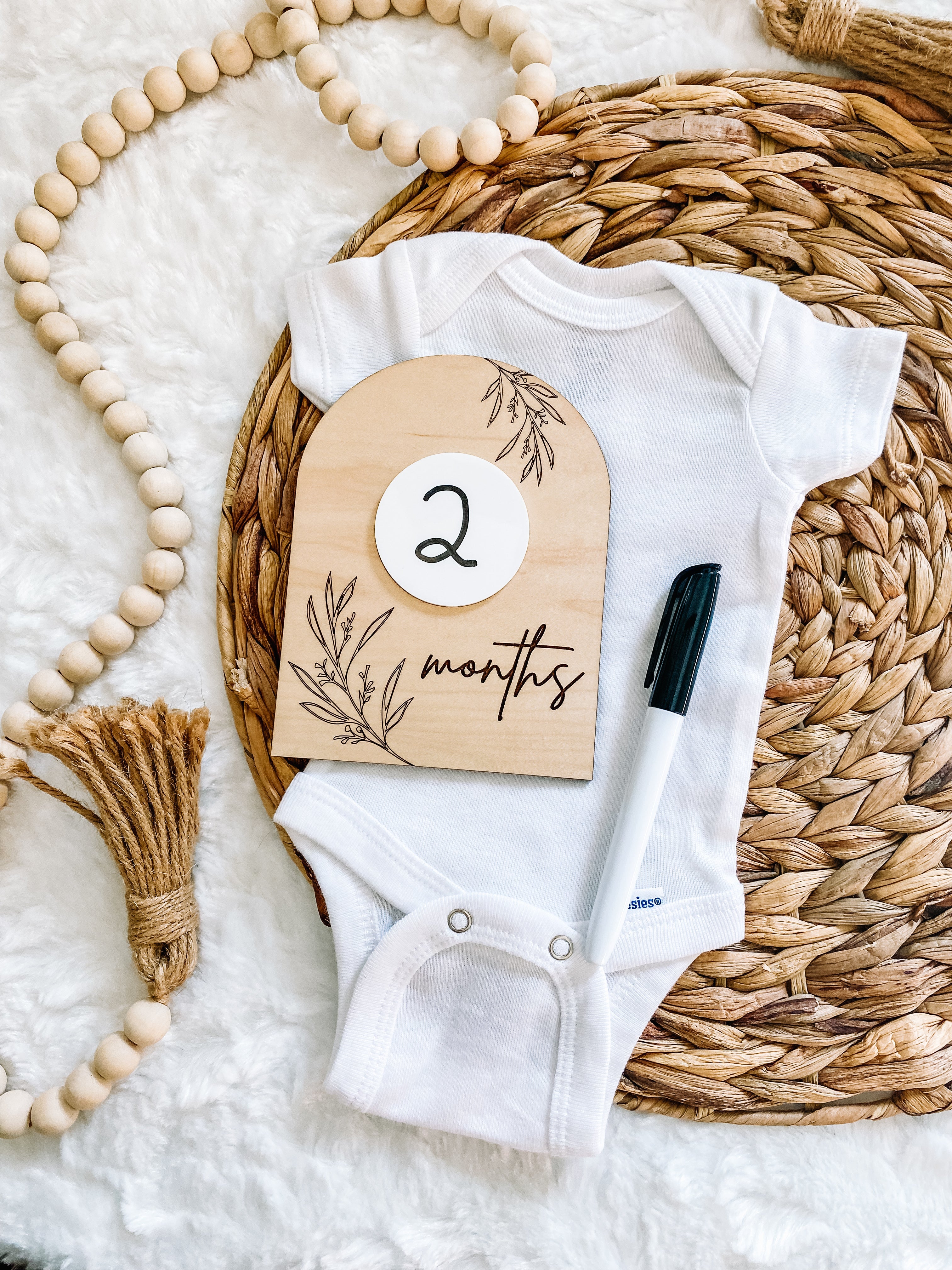 White baby onesie with 2 months milestone card on woven mat, ideal for baby clothing and keepsakes.