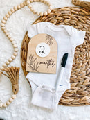 White baby onesie with 2 months milestone card on woven mat, ideal for baby clothing and keepsakes.