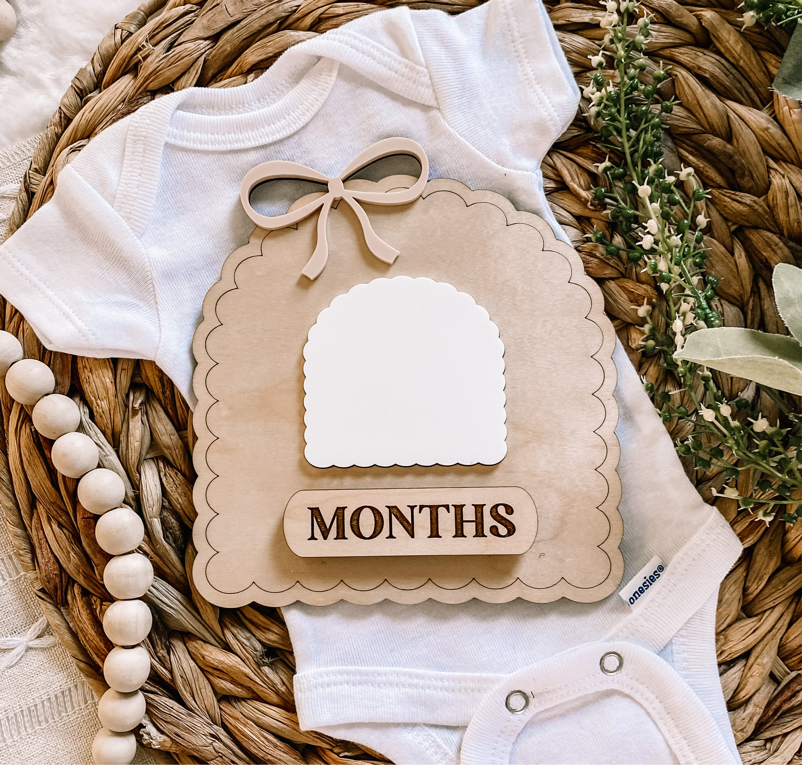 White baby onesie with wooden 'MONTHS' milestone sign, bow, and beads on woven mat