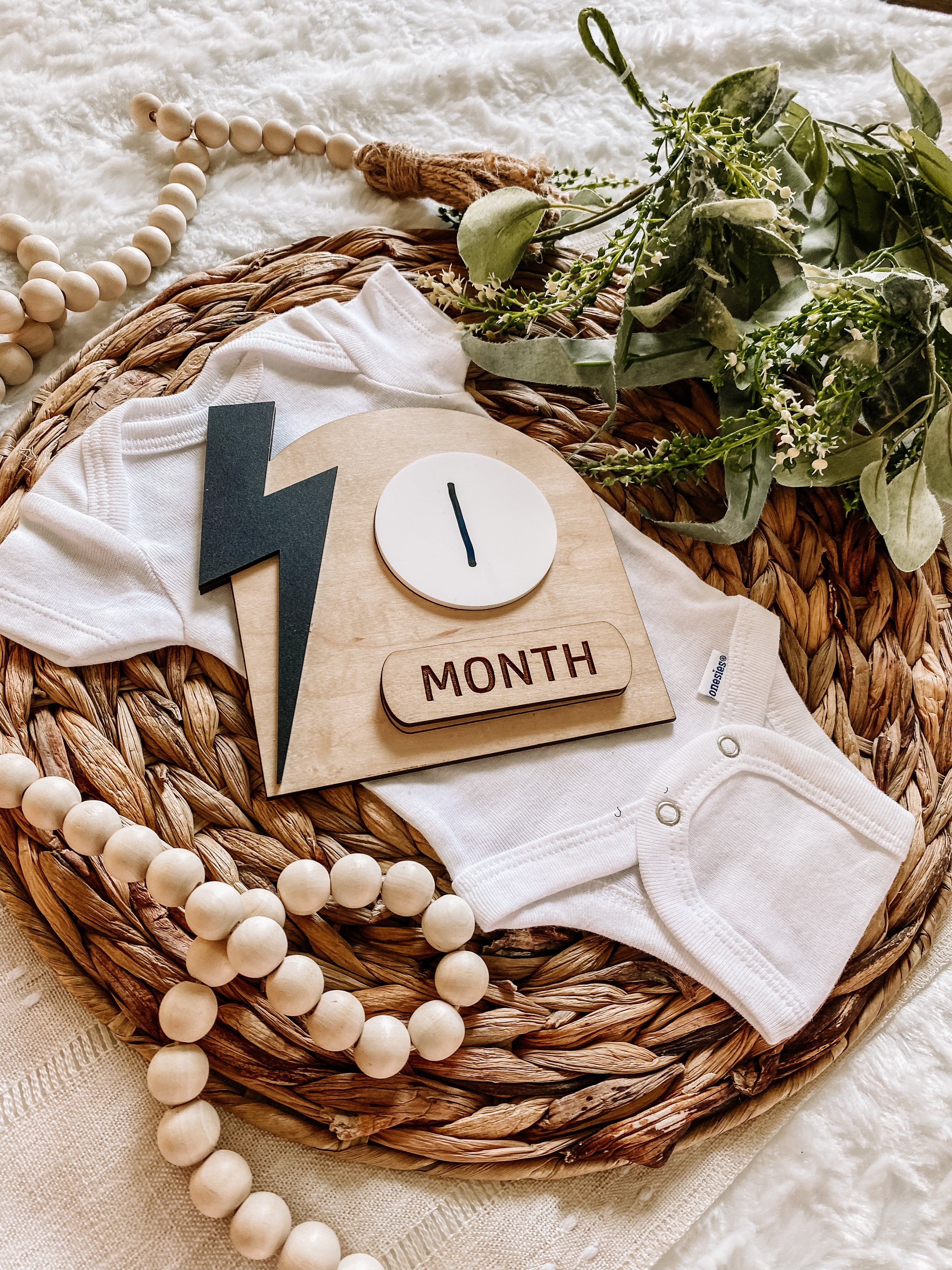 White baby onesie with wooden '1 Month' milestone plaque, greenery, and beads on woven mat