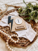 White baby onesie with wooden '1 Month' milestone plaque, greenery, and beads on woven mat