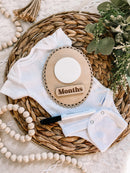 White baby onesie with wooden months milestone plaque on woven basket, ideal for baby photo props