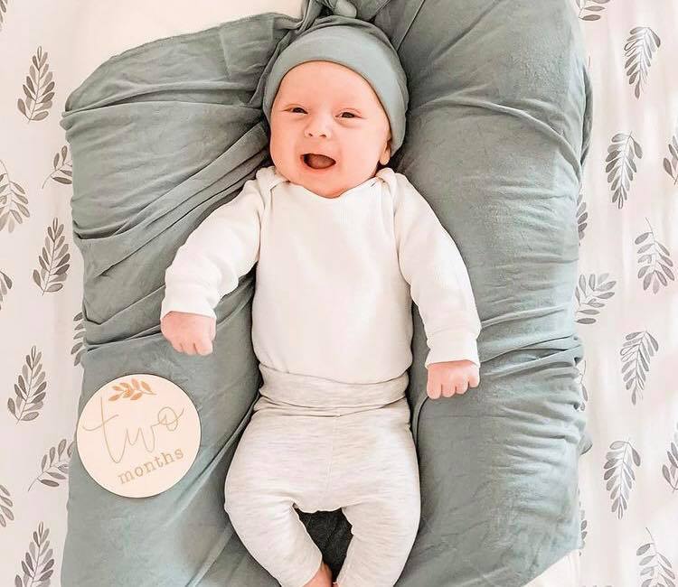 Happy baby in white onesie and gray swaddle, smiling on leaf-patterned blanket with 'two months' sign