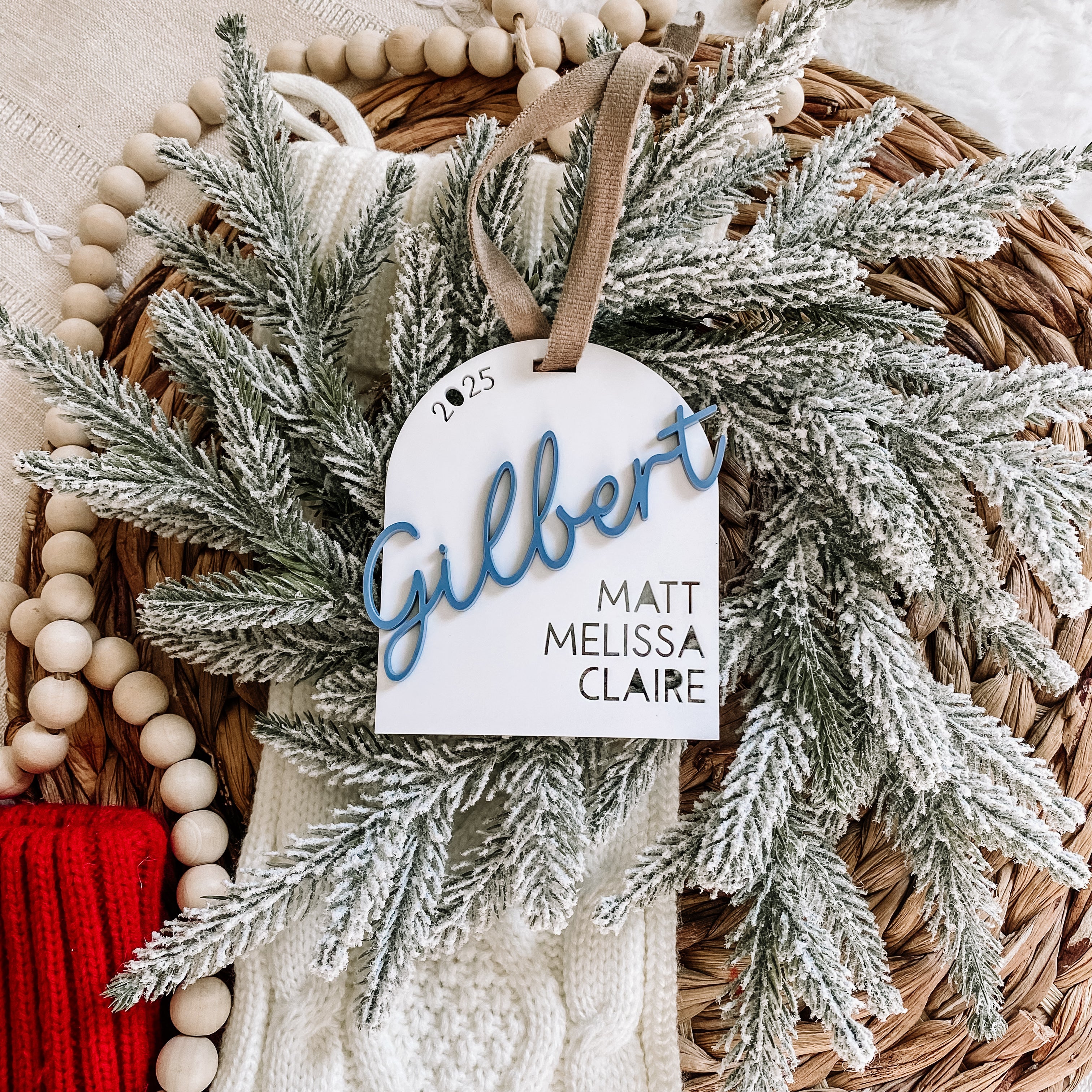 Personalized 2025 family ornament with 'Gilbert' in blue script, nestled in frosted greenery and wooden beads, perfect for holiday decor.