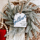 Personalized 2025 family ornament with 'Gilbert' in blue script, nestled in frosted greenery and wooden beads, perfect for holiday decor.