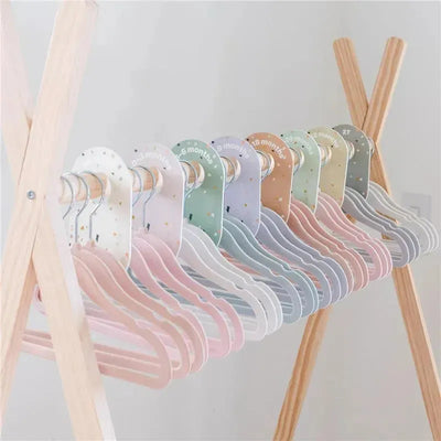 Velvet baby hangers in pastel colors, labeled with size indicators, displayed on a wooden rack. Pack of 15, non-slip and durable.